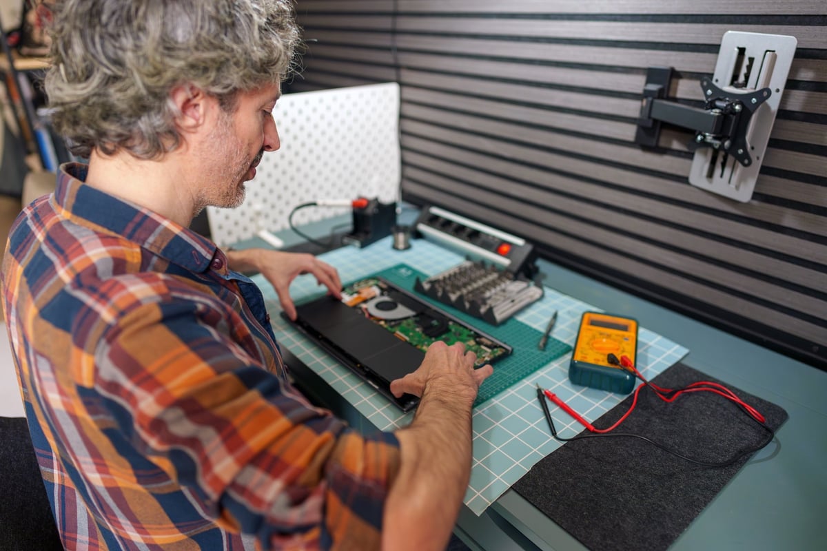 Male electronics technician repairing an open laptop, using tools like a multimeter and screwdrivers on a workbench
