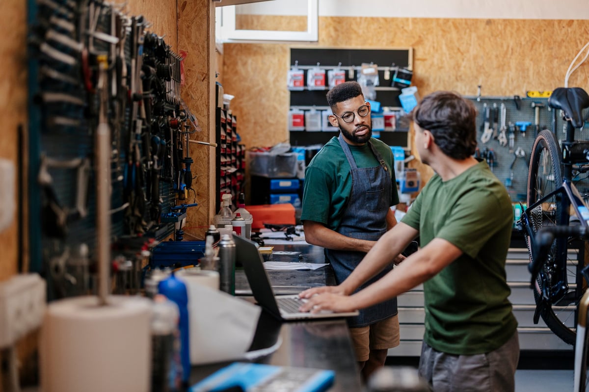 Two men collaborating in a busy bicycle repair shop. Discussing service details while one works on a laptop next to tools and equipment. Representing small business operations and customer interaction