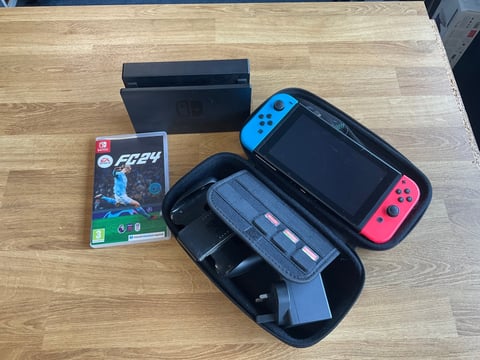 Nintendo Switch gaming console with blue and red controllers displayed with carrying case and FIFA 24 game on wooden surface